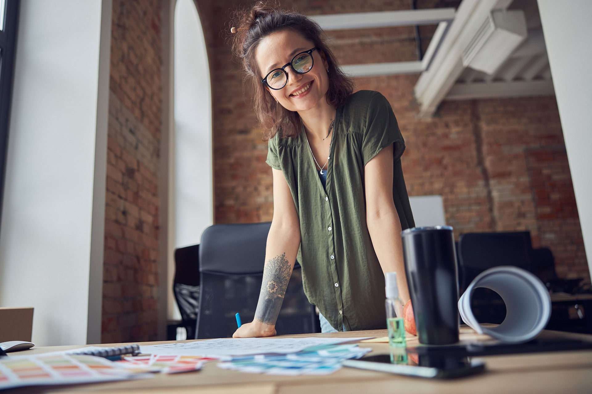 Creative woman, interior designer or architect in casual wear with messy hairdo smiling at camera while working on a blueprint for new project, standing in her office